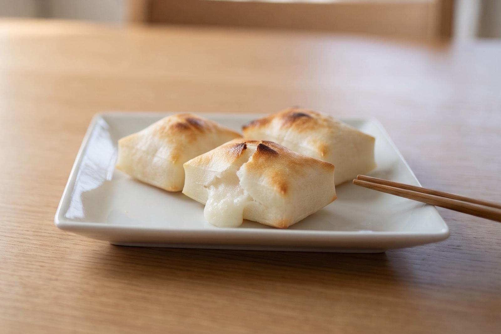 木のテーブルの白い角皿に、焼き目がついてふくらんだ切り餅が3個。手前の1個は裂け目から白いお餅がはみ出している