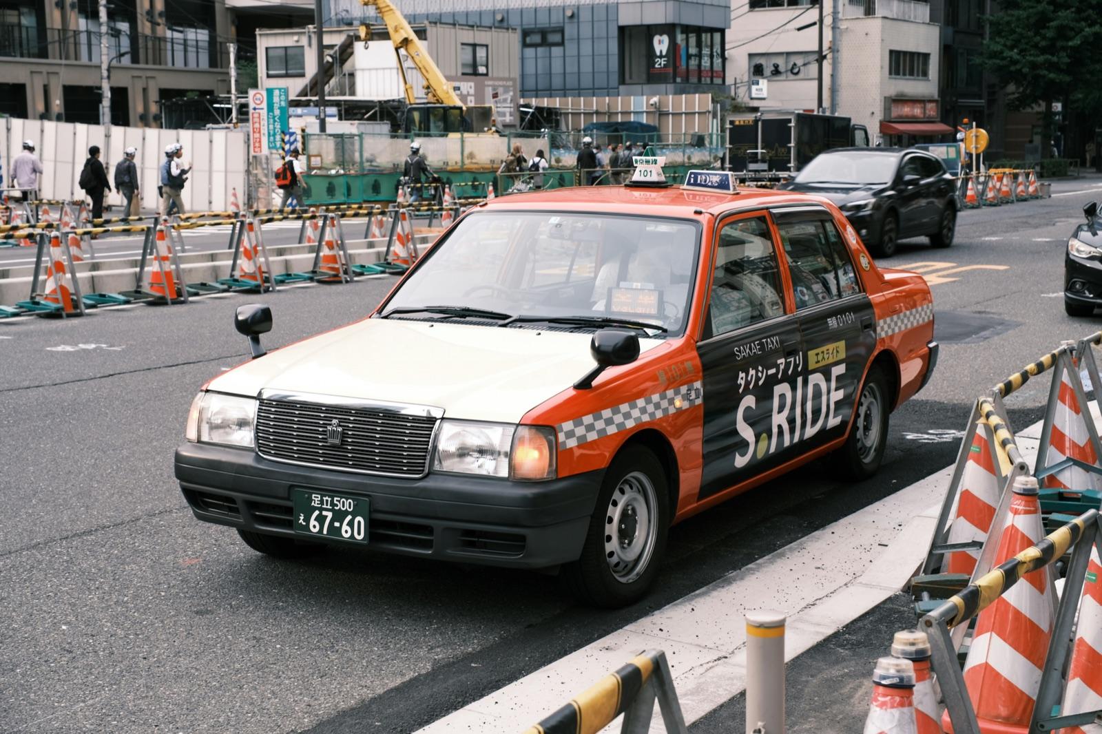 工事の柵が並ぶ街なかの道に出てきた、オレンジとクリーム色の車体の東京のタクシー