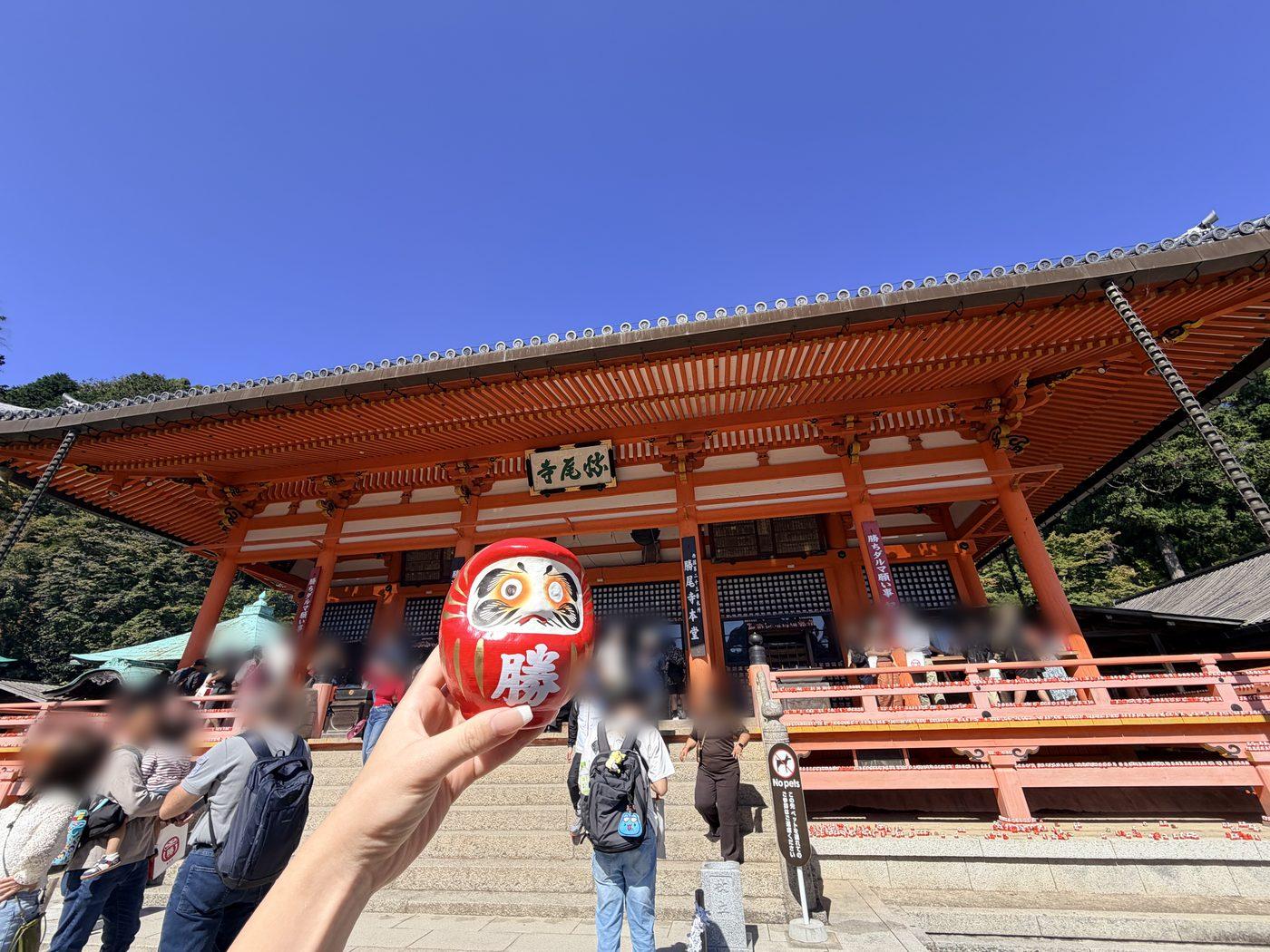 朱色の本堂を背に、手に持った小さな赤い勝ちダルマ。「勝」の字が書かれている