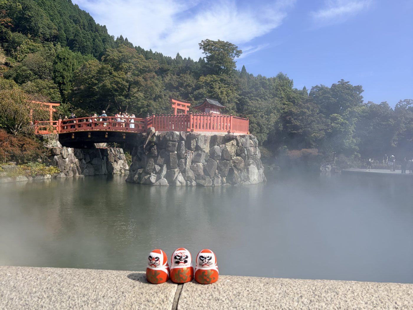 境内の池に朱色の橋と小さなお堂が立ち、後ろに緑の山が広がる。手前の石の手すりの上に、小さな赤いだるまが3つ並んでいる