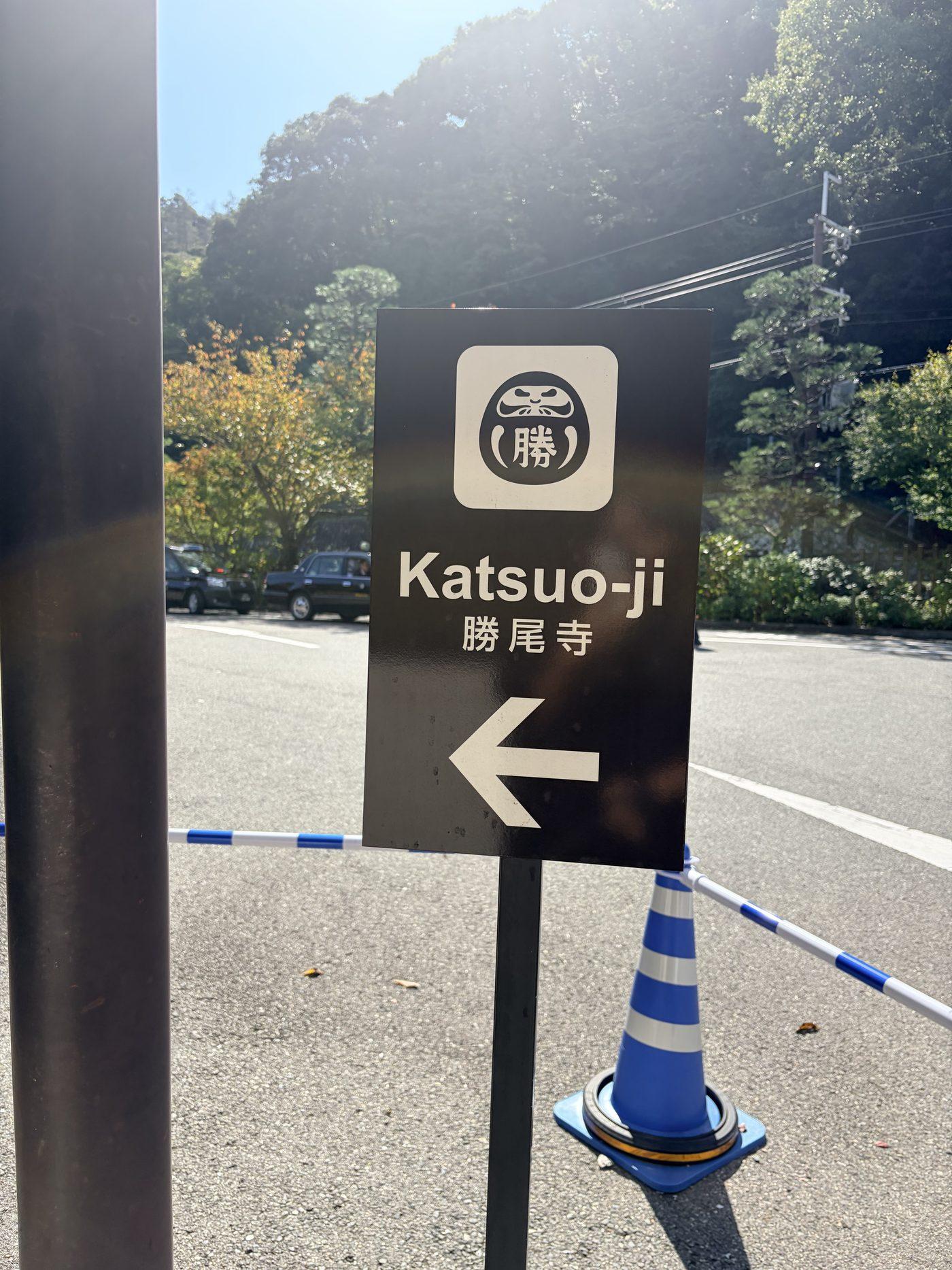 舗装された駐車場に立つ、勝尾寺のだるまのマークと「Katsuo-ji 勝尾寺」の文字、左向きの矢印の案内板
