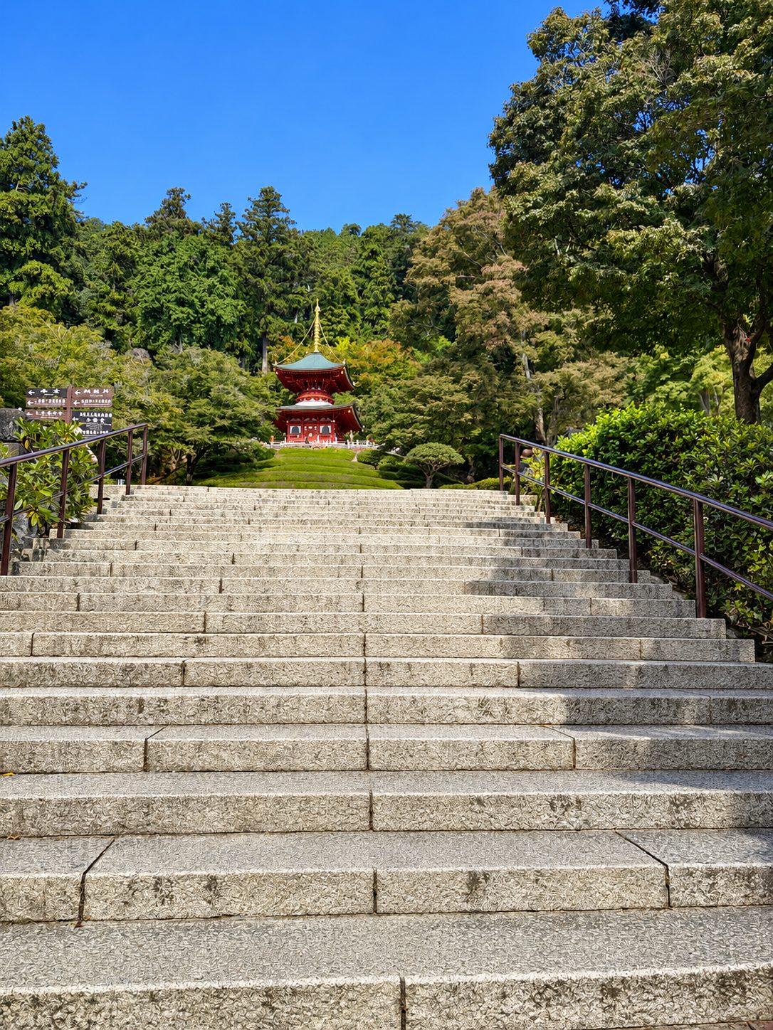 手すりのついた長い石段がまっすぐ上へ続き、その先の緑の木々の中に朱色の多宝塔が立っている