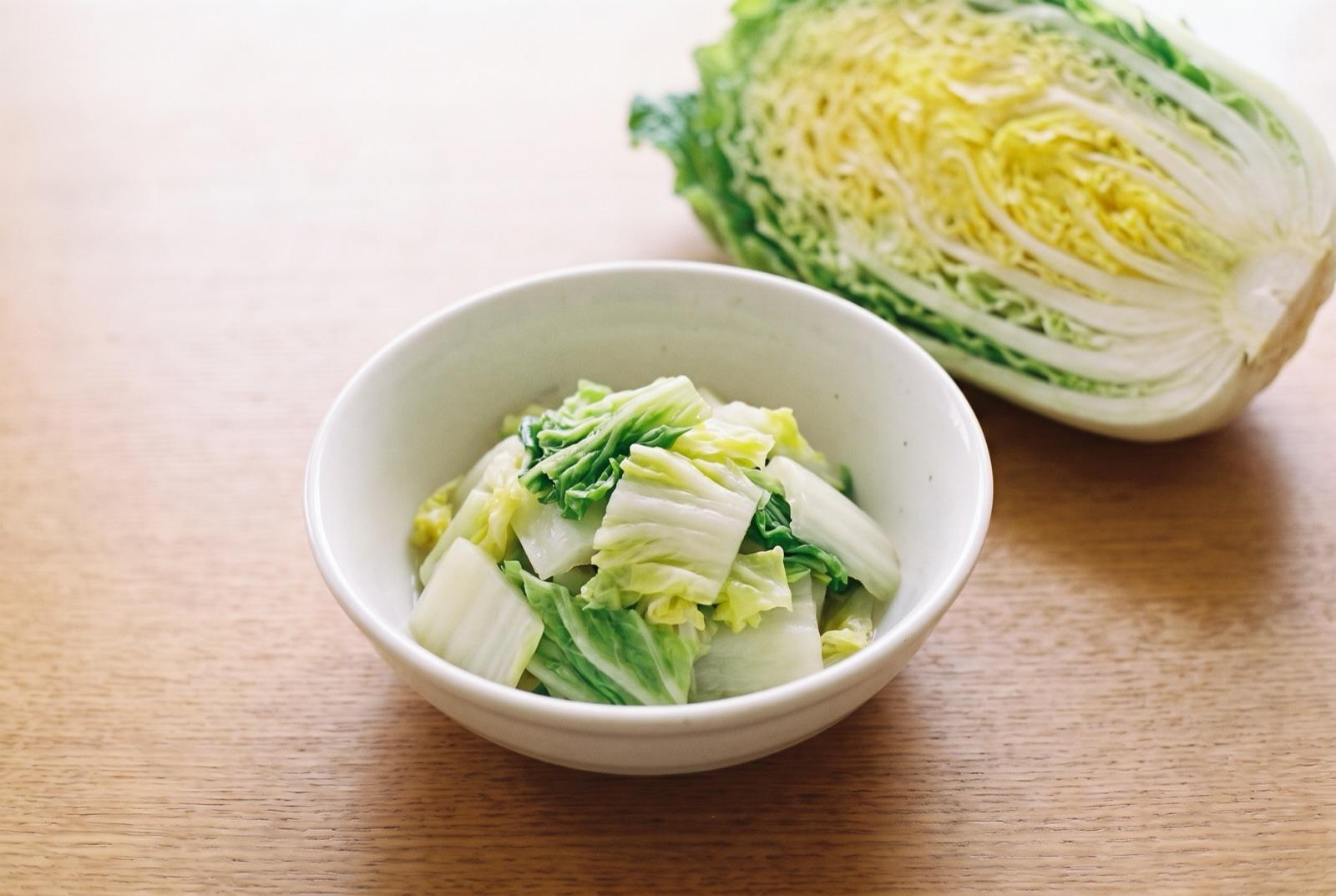 明るい木のテーブルの白い器に、ゆでて刻んだ白菜。奥に半分に切った白菜