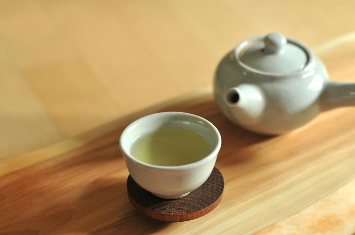 木のテーブルの上の、木のコースターにのせた白い湯のみのせん茶と、奥の白い急須