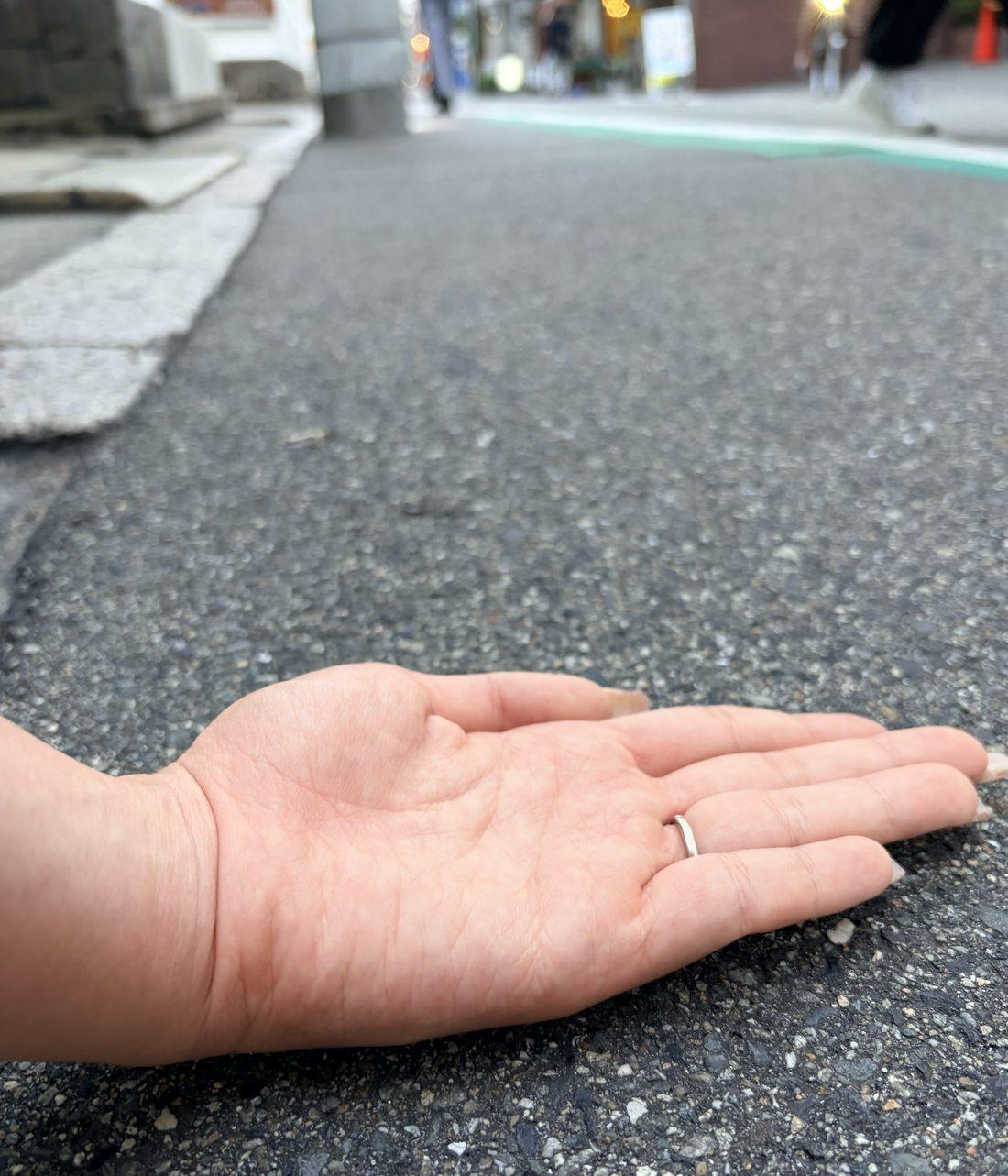 道路のアスファルトに手の甲をあてて、路面の熱さを確かめている手元。奥には夕暮れの街並みがぼやけて見える